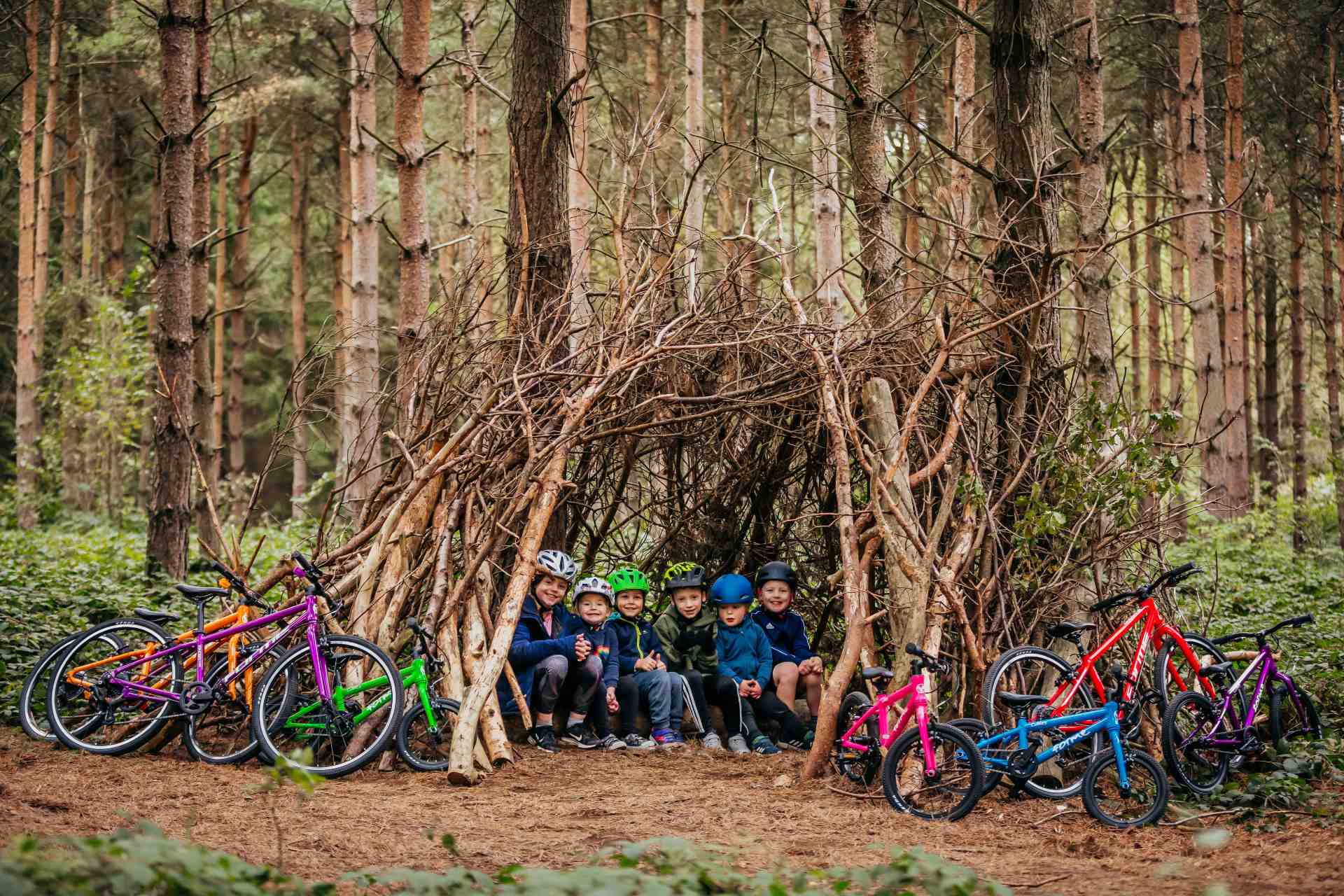 A group of young cyclists gather together in the forest in a den made out of branches with their Forme Cubley and Kinder bikes nearby.