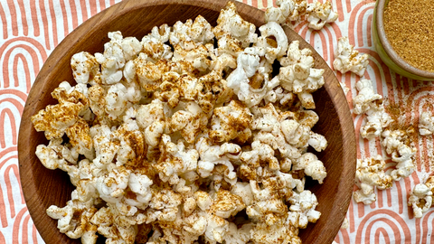 A wooden bowl of popcorn tossed in organic spices, Firecracker Dust from Well seasoned table, and a bowl of seasoning blend on the side on a textile background.