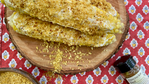 A wooden platter of elote, Mexican street corn, made with Firecracker Dust from Well Seasoned Table, on a textile background with a glass shaker jar of the organic seasoning.