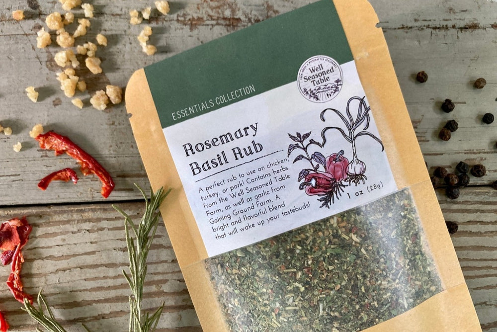 A packet of Rosemary Basil Rub from Well Seasoned Table on a wooden background with peppers, garlic, and rosemary.