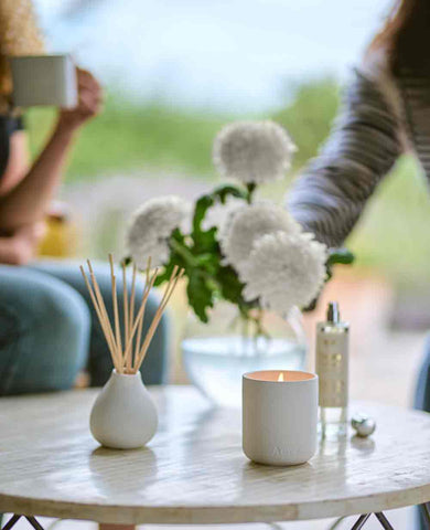 two people next to coffee table with lit candle diffuser and room mist along with a bunch of flowers