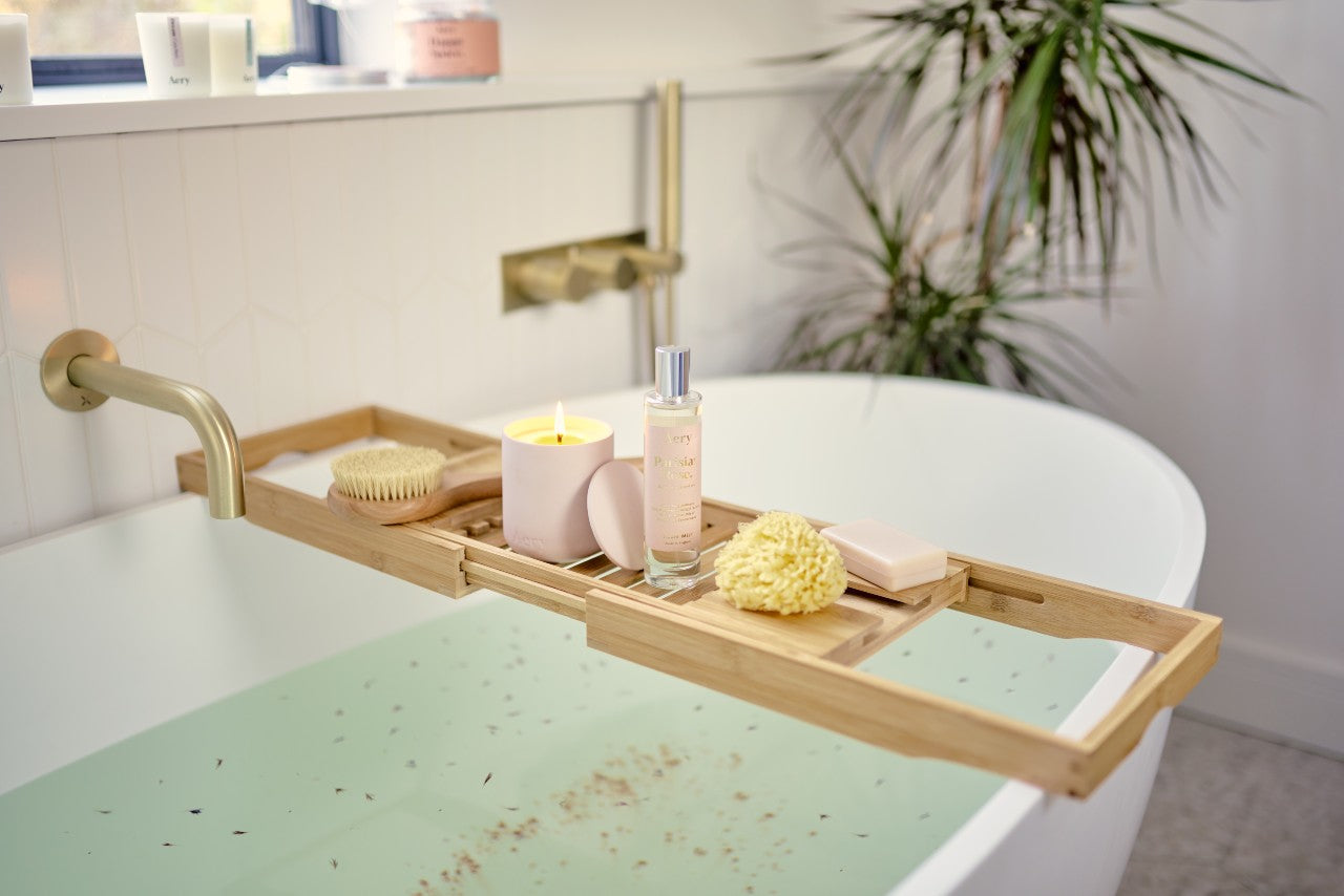 Run bath filled with water. A wooden bath tray filled with bath accessories, a lit candle and other Parisian Rose products lays across the bath. There is a plant pot in background and natural light from window.