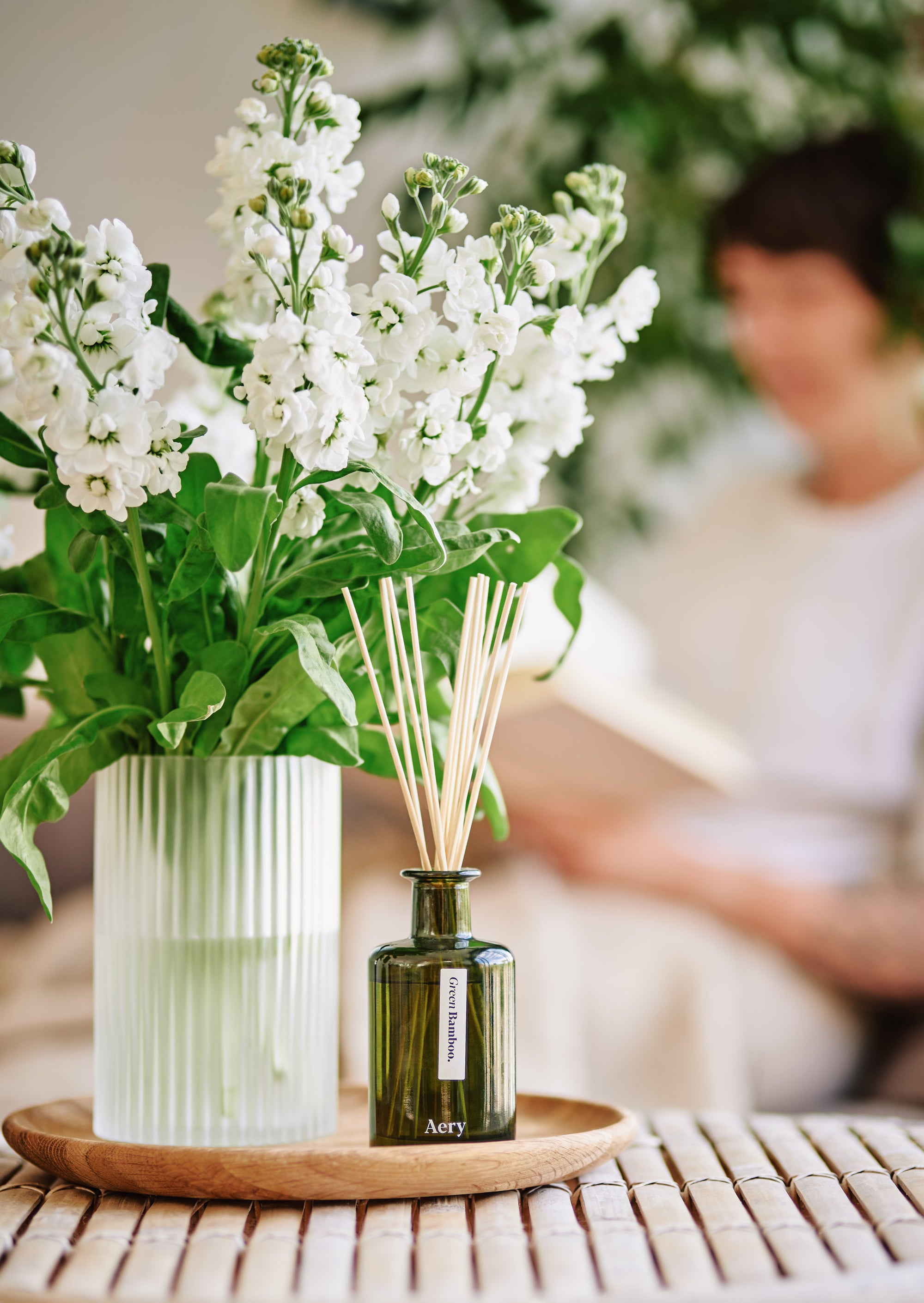 coffee table setting with tray and vase of white flowers next to an aery living reed diffuser in foreground and person sitting reading in the background 