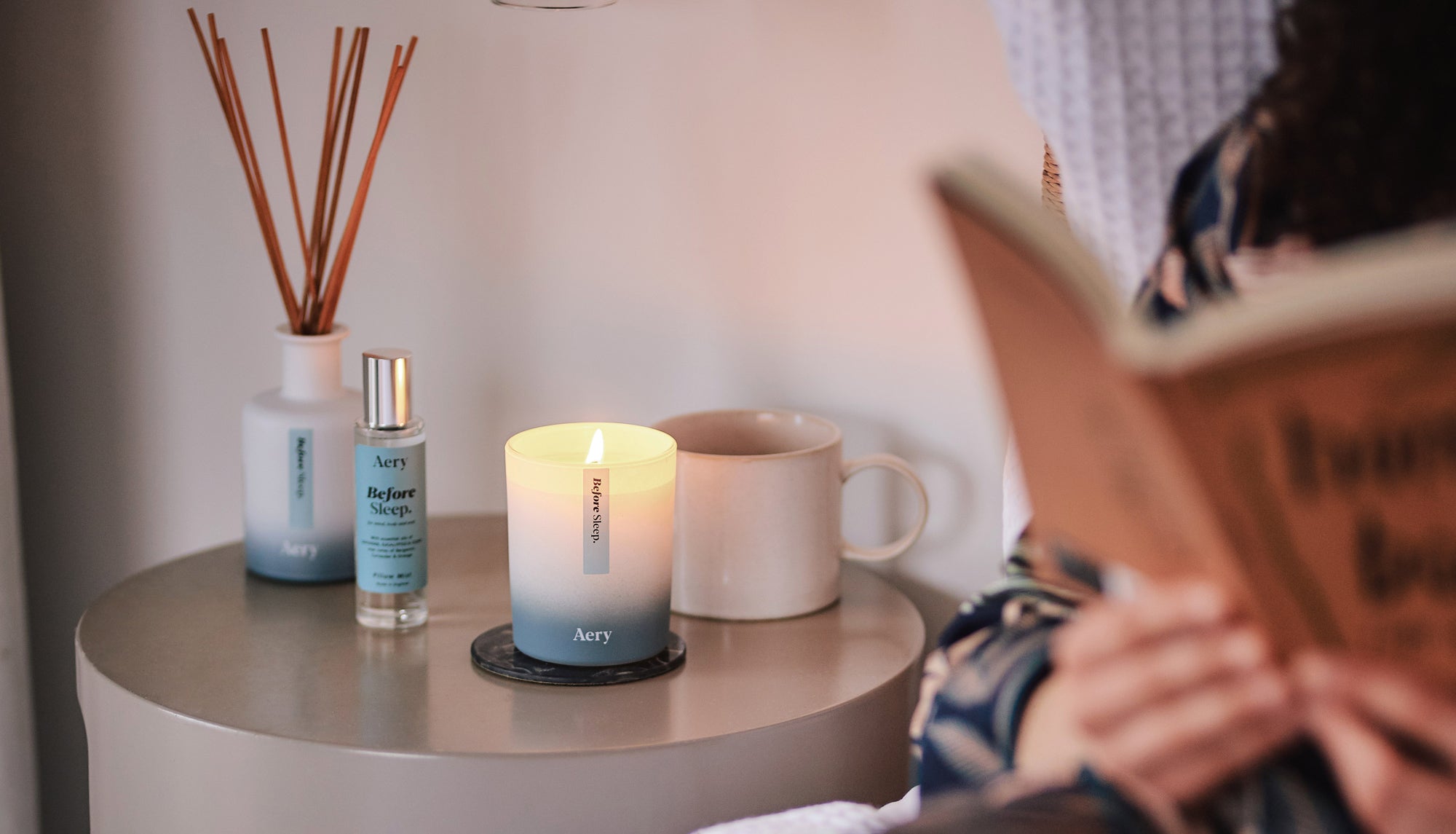 person reading in bed with aery before sleep diffuser, candle and cup of tea on bedside table