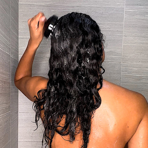 a dark complected woman washes her brown curly in the shower with the shampoo brush