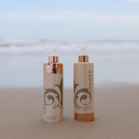 cream and gold colored shampoo and conditioner bottles sit on a beach