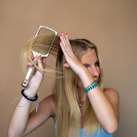 a blonde woman brushes through her hair with a golden colored paddle hair brush