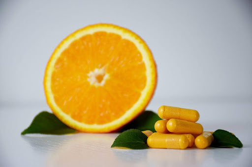 A slice of orange standing next to orange vitamin C supplements against a gray background, with green leaves underneath them.