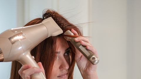 a woman with red hair using a triangle shaped brush and blowdryer to create dramatic volume near the roots of her hair.