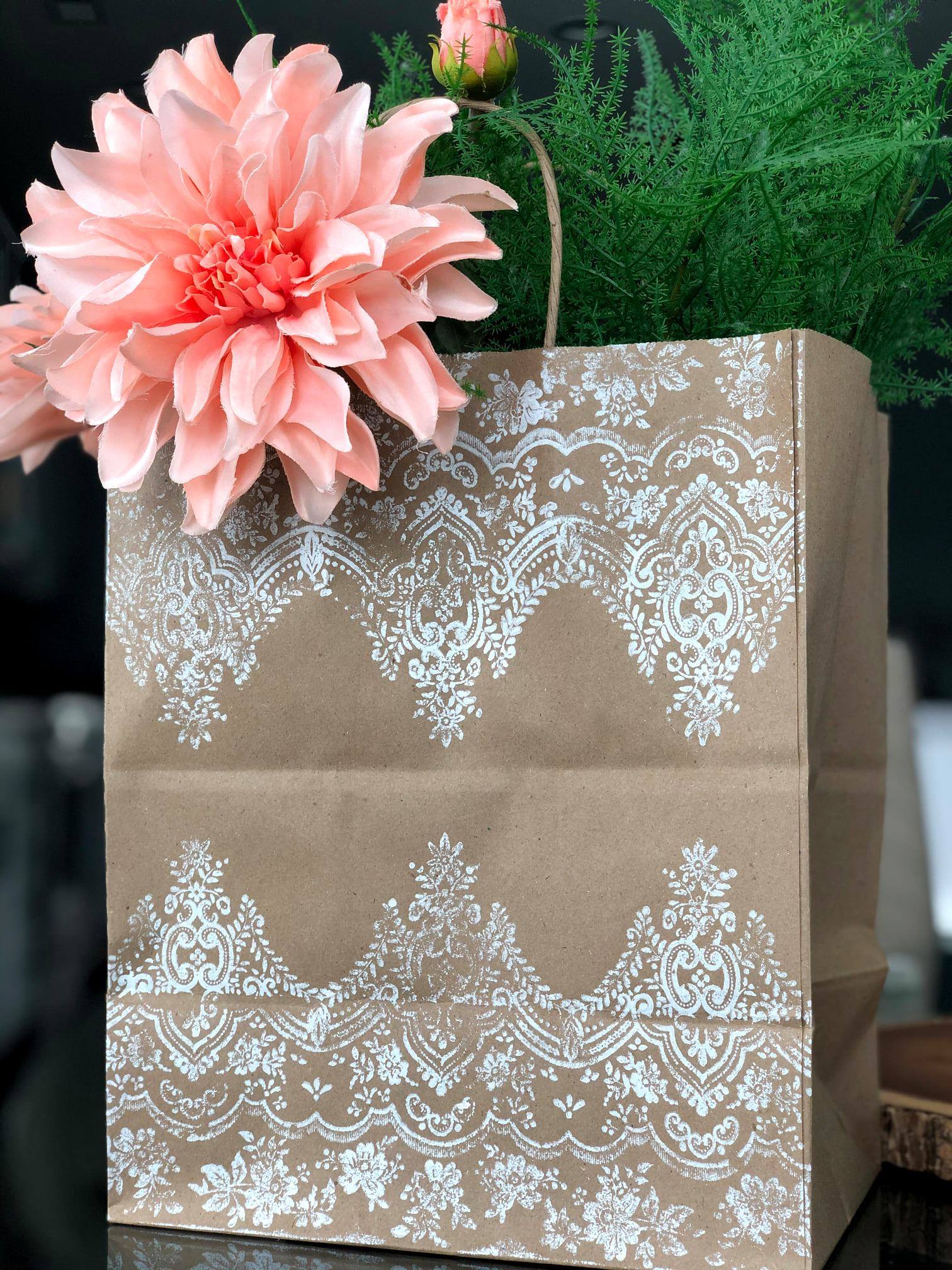 A Kraft bag with a white stamped pattern on the outside. Sticking out of the bag is a large pink flower and some greens.