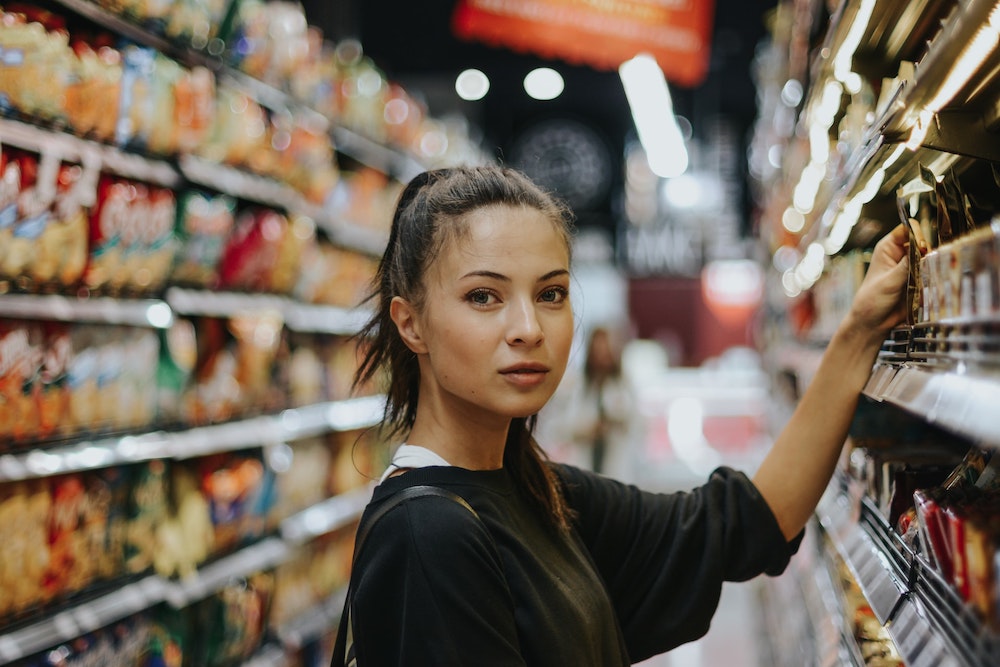 Chica joven mirando de frente y cogiendo un artículo de la estantería de un supermercado