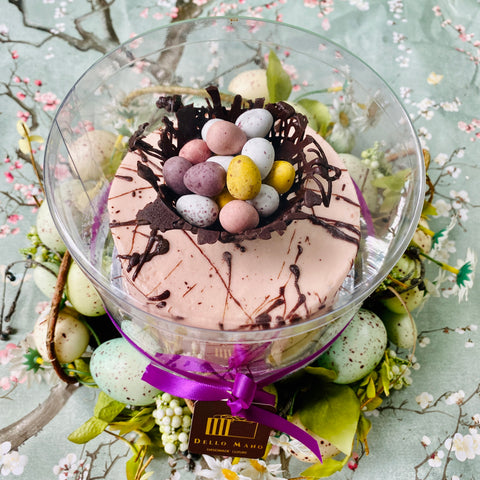 The Dello Mano Easter Egg Cake gift from above you can see pink frosting, chocolate speckles and a chocolate nest holding mini Easter eggs. The cake stand is surrounded by green Easter Eggs.