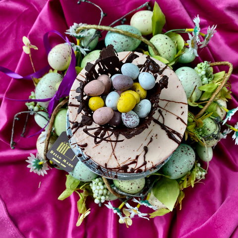 Top view of the Dello Mano Easter Egg cake shows the pink frosted cake with a chocolate nest full of Eater eggs. The cake stand is surrounded by Easter eggs.