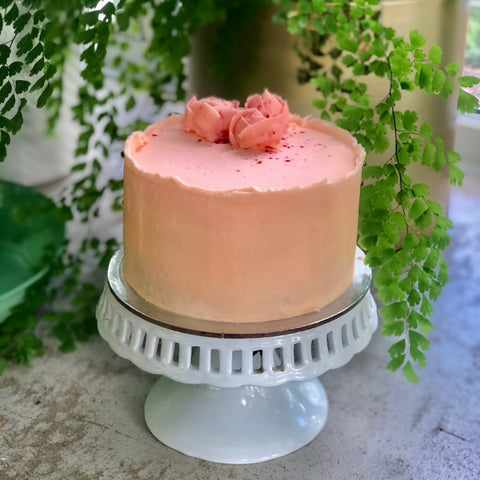 A pink Mothers Day Rose Cake on a white stand with greenery behind.