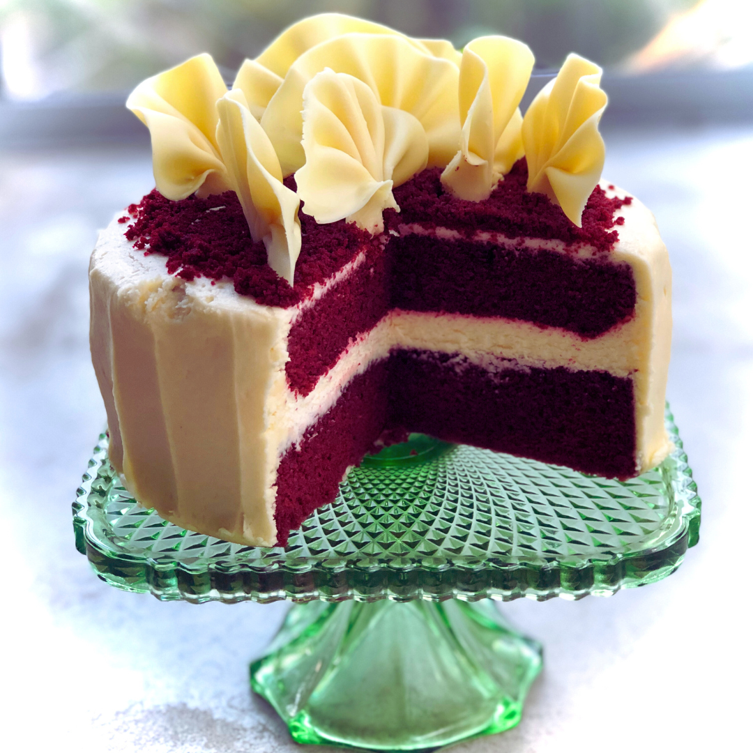 A Dello Mano Red Velvet Cake with red flowers and a green vase and pears in the background