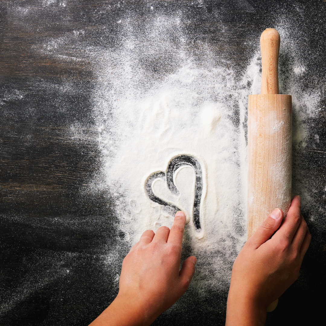 a  hand making a heart shape in flour with rolling pin at side