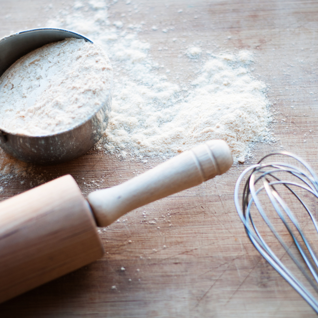 a cup of flour, rolling pin and whisk on a wooden bench