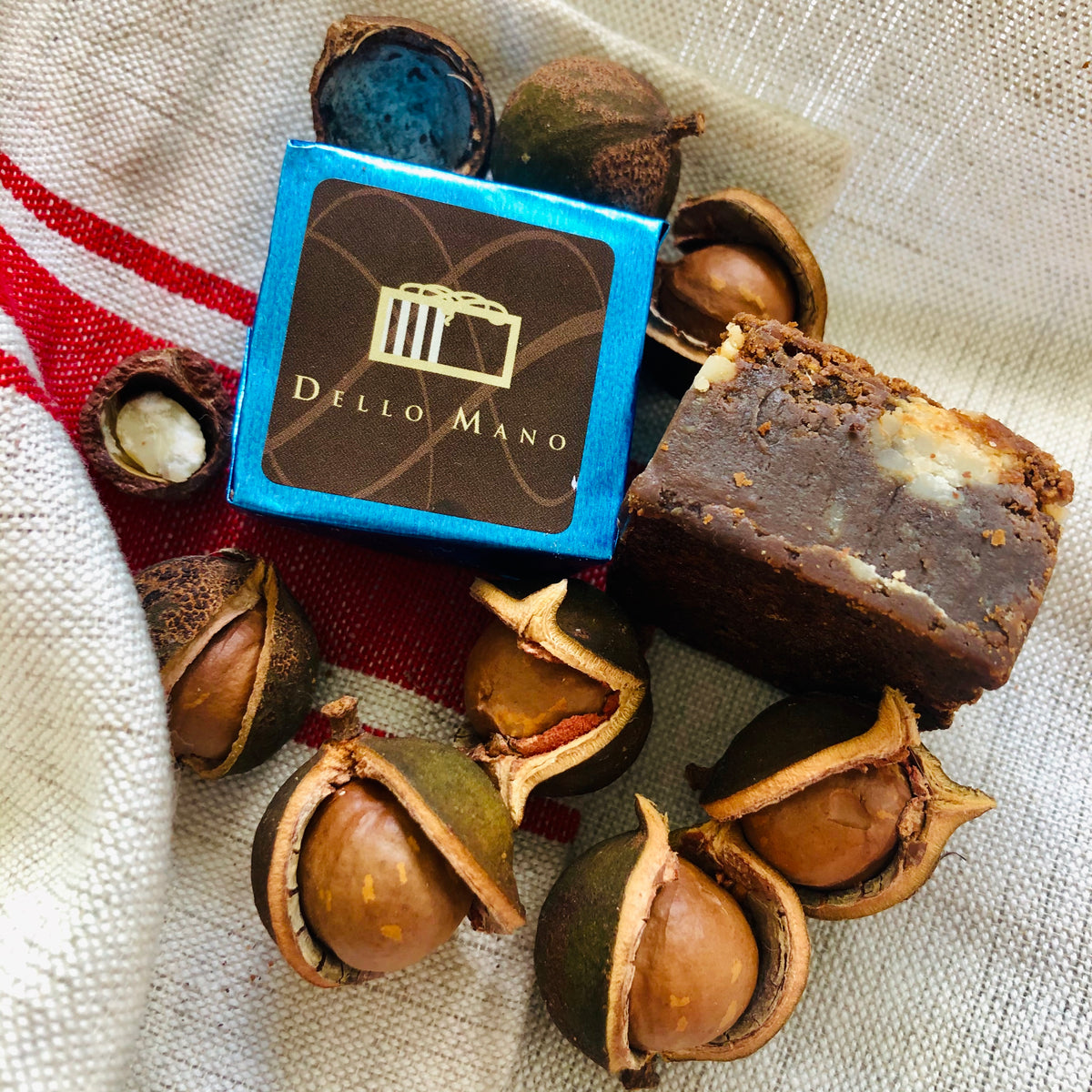 A blue foiled honey caramelised macadamia brownie with one open beside it and surrounded by macadamia nuts in thier shells