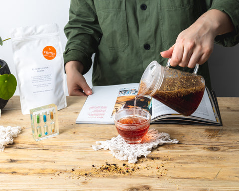 Baies de fleurs de sureau versées dans une tasse par quelqu'un lisant un livre avec des feuilles de thé en vrac sur le comptoir. Une minuterie à thé sur le côté et un sac de vente au détail exposé.