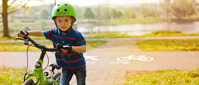 Boy pushing his bike on a bike trail near a lake while wearing a green Melon Helmet