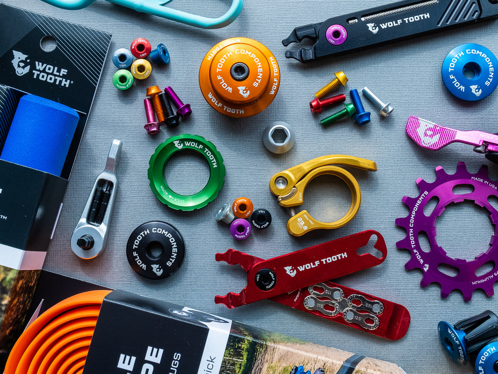 A collection of colorful Wolf Tooth components, including Supple Bar Tape, Seatpost Clamps, Water Bottle Cage Bolts, Grips, Singlespeed Cogs, and Stem Caps.