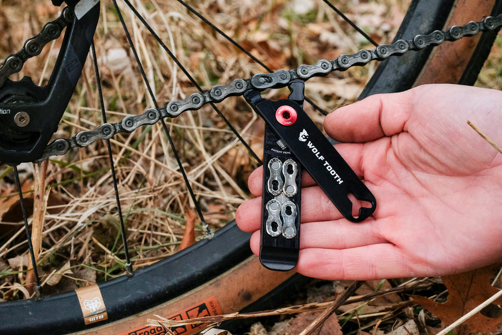 A pair of Wolf Tooth Pack Pliers are held by a cyclist. A bike's chain is in view. The cyclist is using the Wolf Tooth Pack Pliers to break or adjust the bike's chain.