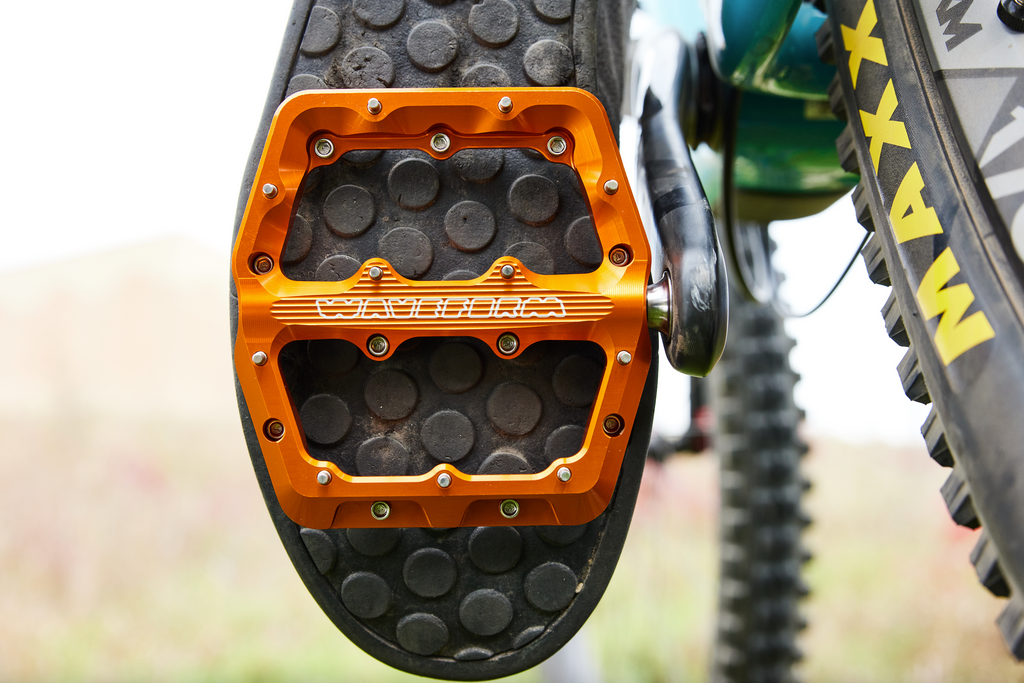 A shot of a cyclist mid-ride. The camera is close up on the rider's foot, showing the detail of the new Wolf Tooth Waveform Aluminum Pedals.