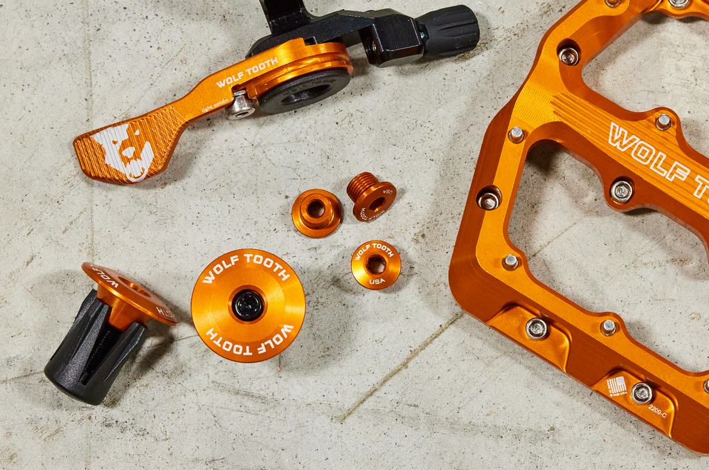 A collection of orange Wolf Tooth products: Orange Waveform Aluminum Pedals, Orange Alloy Bar End Plugs, Orange Chainring Bolts, and an Orange ReMote Light Action.