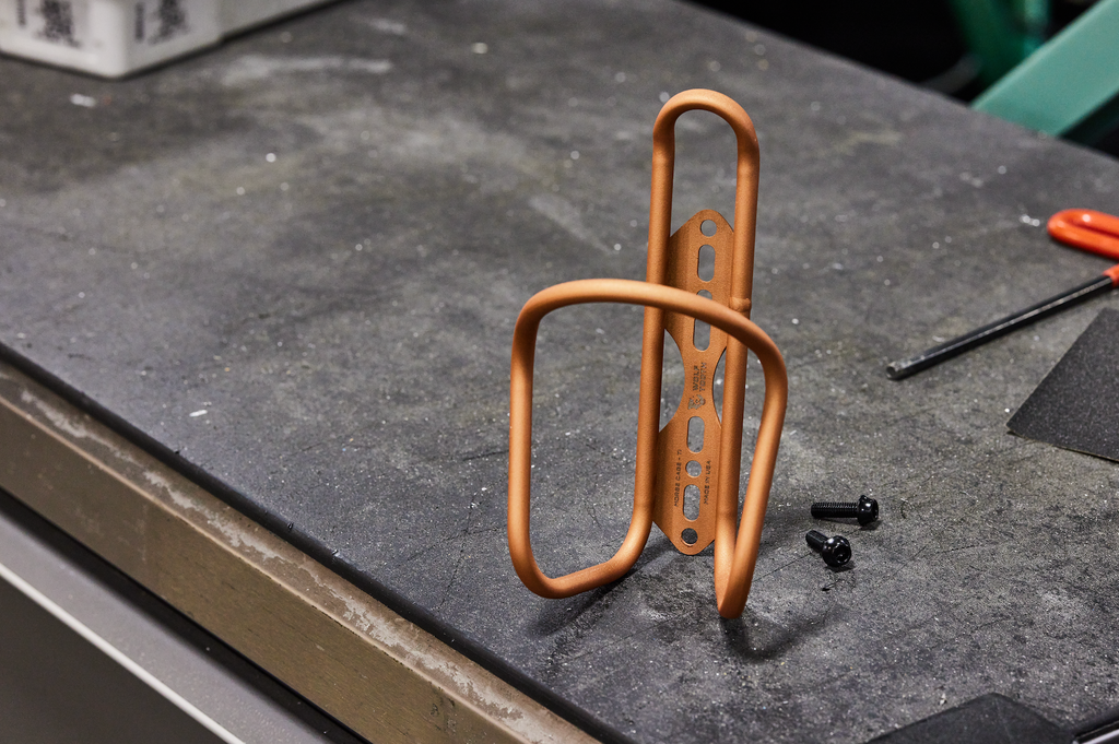 A Wolf Tooth Morse Cage Ti Limited Edition Copper bottle cage sits on a workbench,