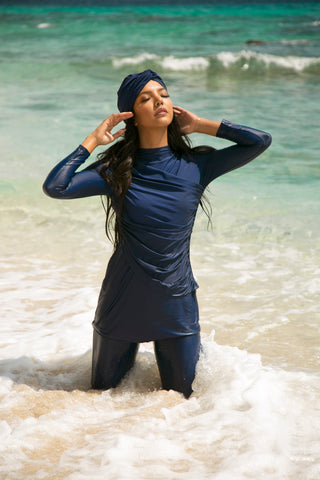 A woman kneeling in the waves of the sea wearing Lanuuk Diana Swimset in Twilight and the Swim Turban in Twilight.