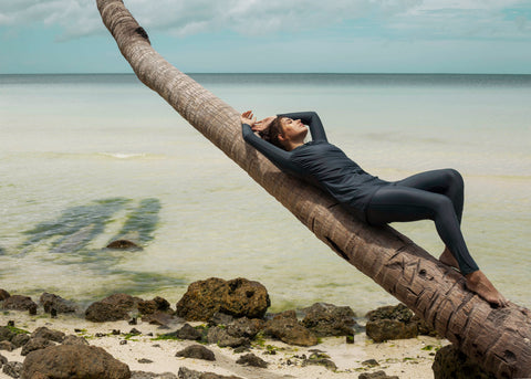 A woman is laying on a palm tree bent sideways in front of the ocean. She is wearing full coverage swimwear in a midnight blue colour.