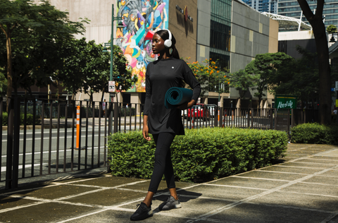 A woman wearing black activewear and white headphones, holding a yoga mat and walking  Caption: Lanuuk Athletic Collection in Black