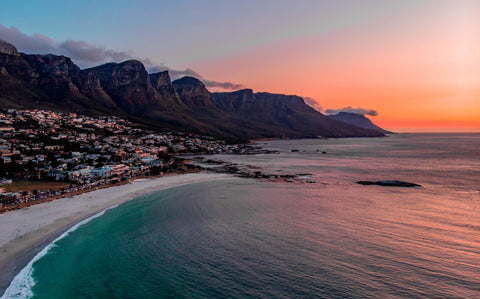 A picture of camps bay in Cape Town, South Africa. The photo is taken via drone and showcases the sun setting over the sea, beach, houses and mountains.