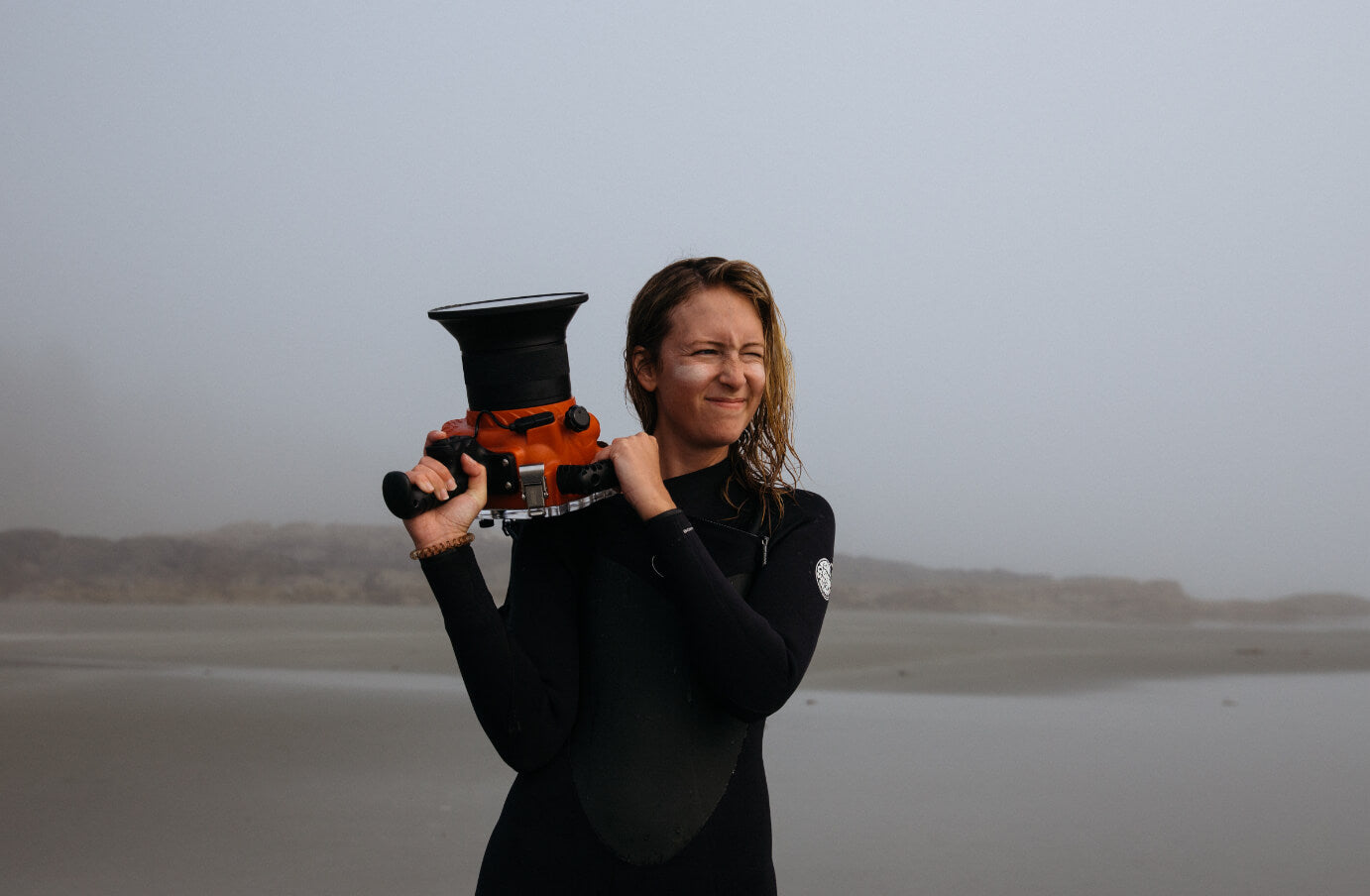Johnie Gall in a wetsuit on the beach holding a camera