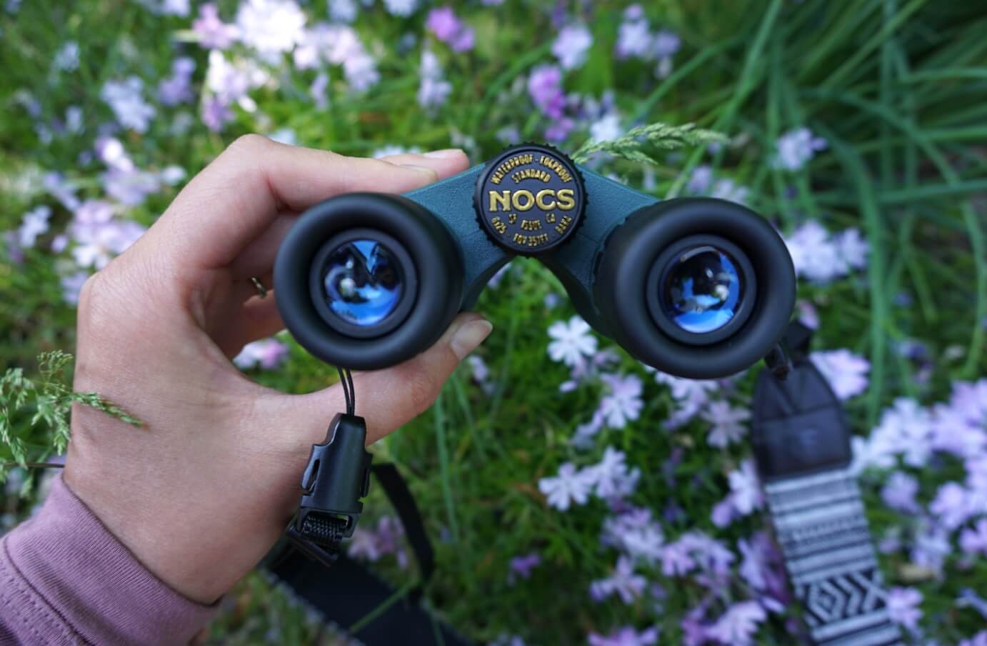 Emily Piper holding a pair of the Cypress green nocs standard issue 8x25 binoculars, looking down towards a green and purple flower bed