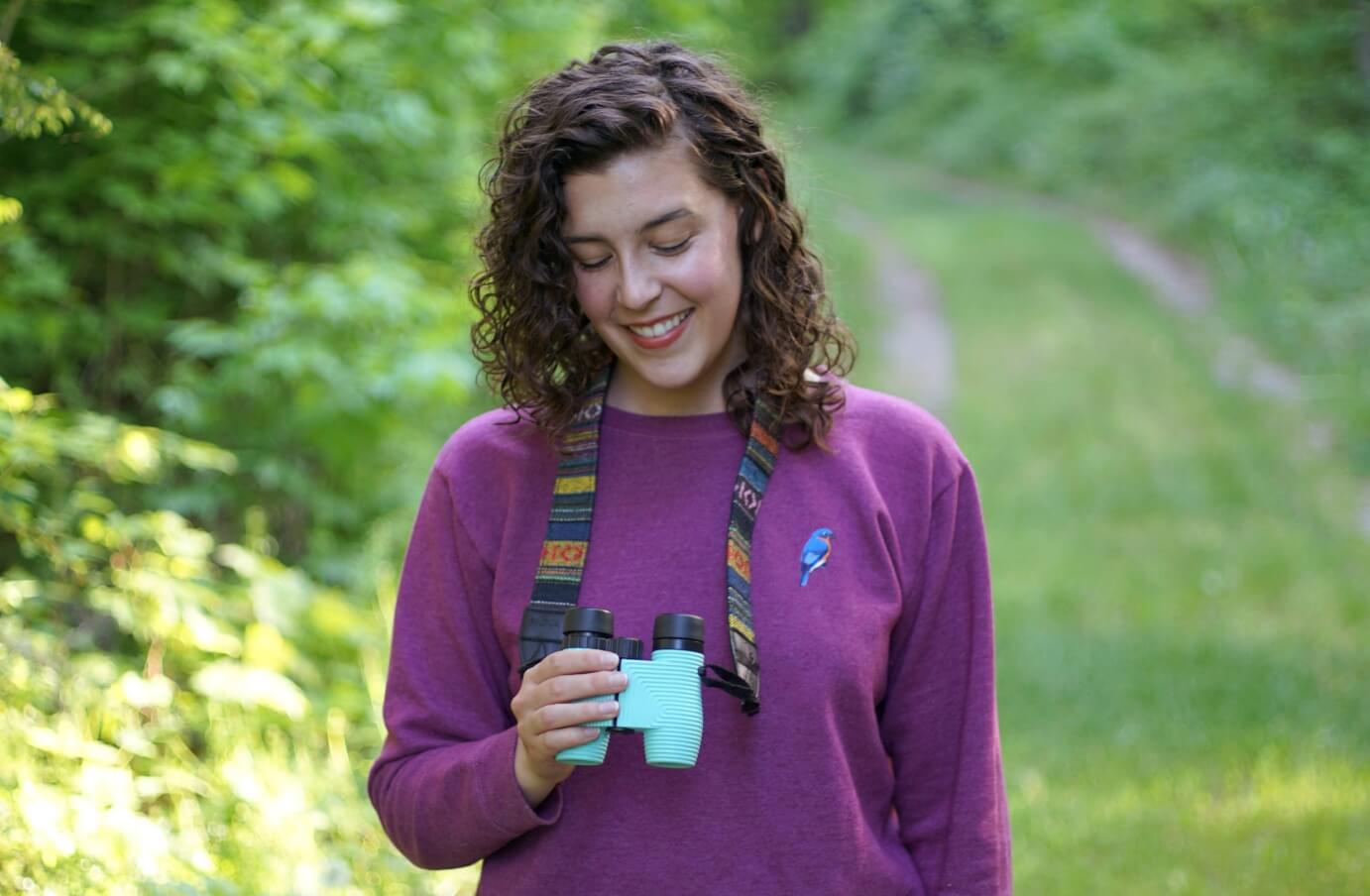 Emily Piper smiling while on a hike surrounded by lush greens, a pair of Nocs standard issue 8x25 around her neck