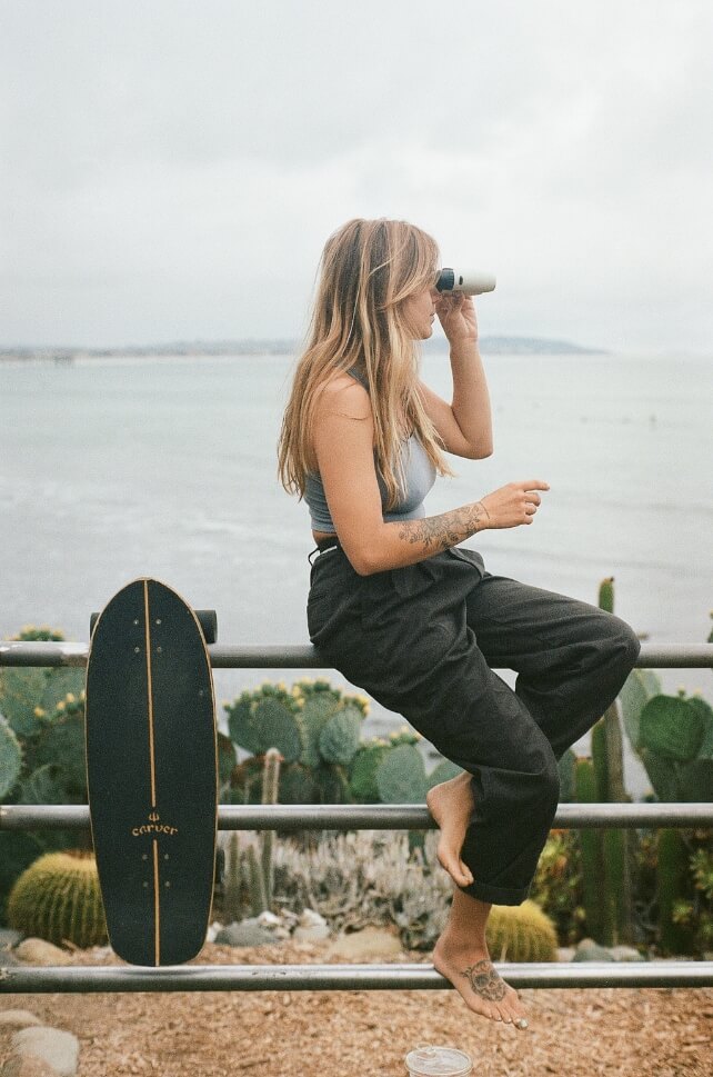 Sarah Rewellc sitting on a fence, looking through a pair of Nocs Standard Issue 8x25 towards the ocean