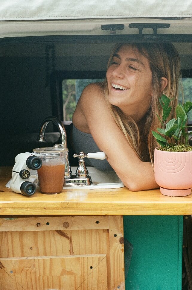 Sarah Rewellc laughing in a camper van, a pair of Nocs Standard Issue 8x25 on the table next to her