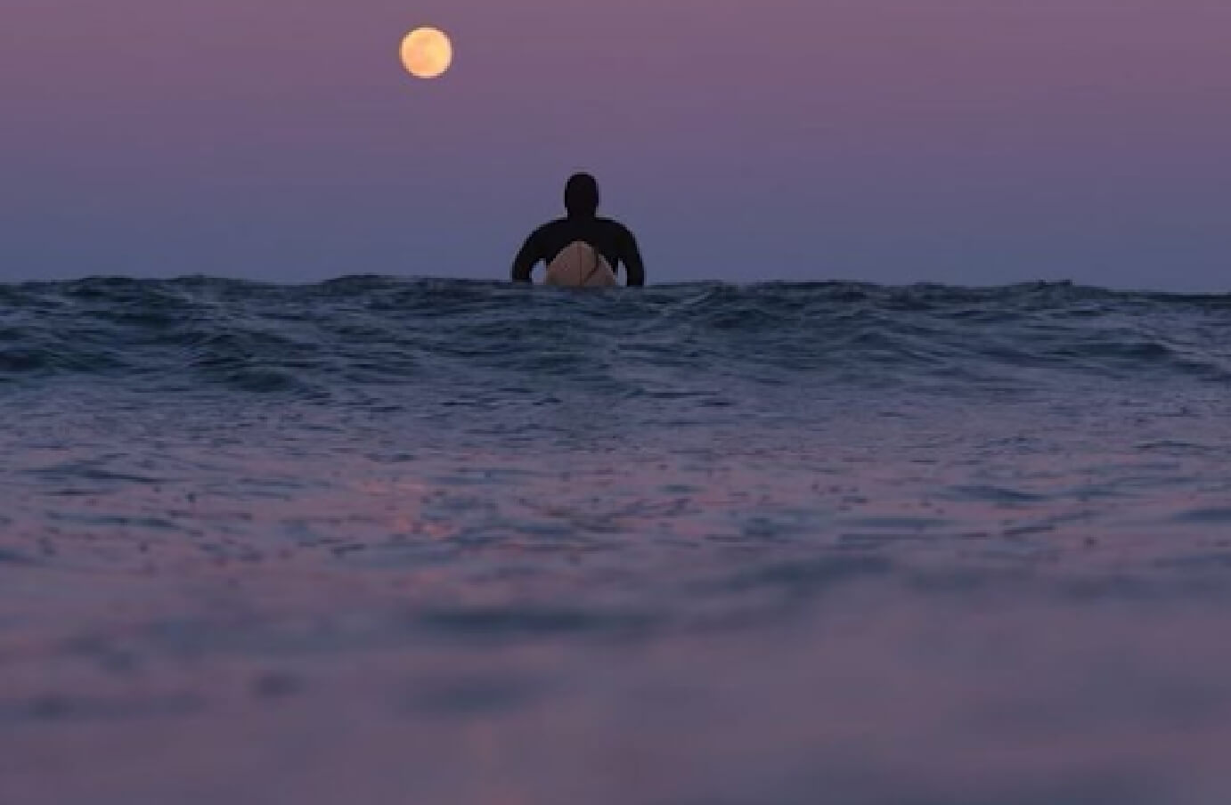 Sunset photography of a srufer on the water, looking out at a moon rising