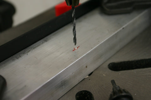 Pilot drill in a drill press about to drill a hole in the bar stock. Intended hole location is marked with red crosshairs.