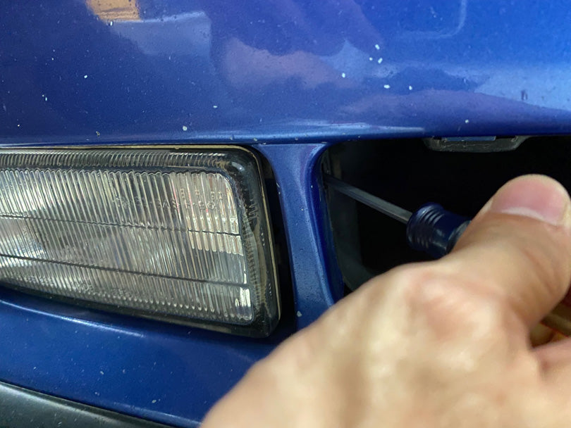 A screwdriver being inserted in a hole in the brake cooling duct to access the release tab that releases the foglight.