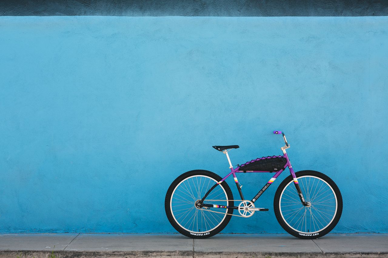 Multi colored bicycle that is Purple, Black and Yellow. Parallel with a bright blue wall  