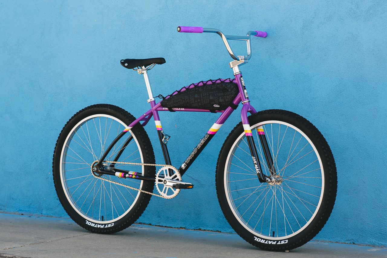 Multi colored bicycle that is Purple, Black and Yellow. Parallel with a bright blue wall  