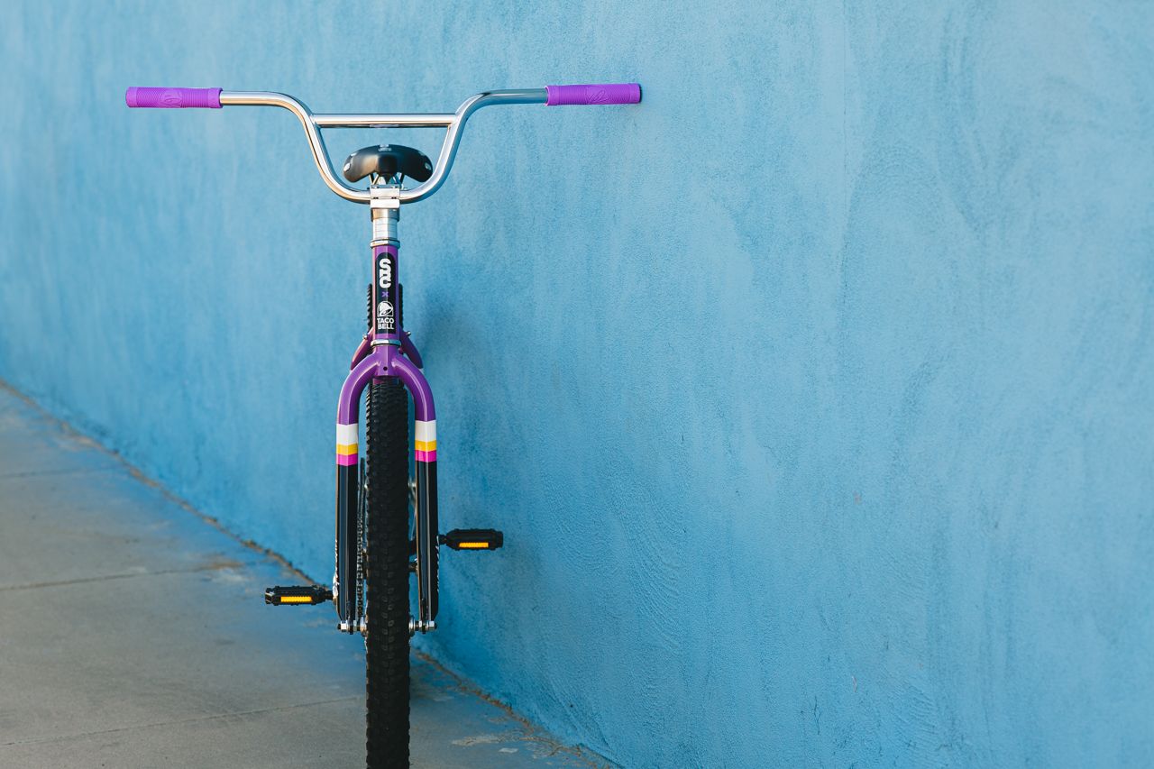 Multi colored bicycle that is Purple, Black and Yellow. Parallel with a bright blue wall  