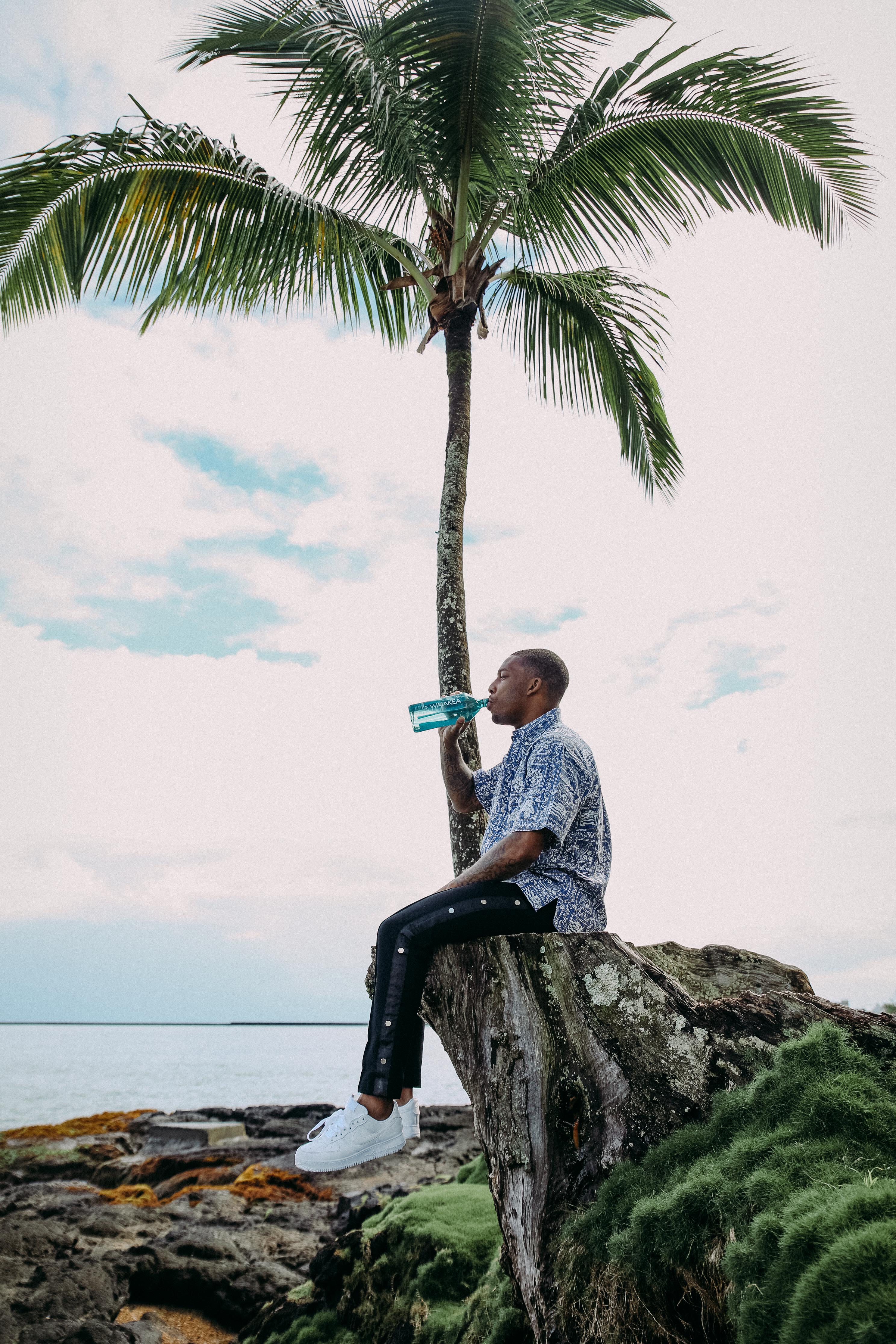 Troy Hill sitting on palm tree drinking Waiākea water Nui 1L Oceanplast 