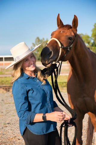 Woman with blond hair wearing a tan nevada style hat, blue button up jean blouse and black May Babes riding breeches laughs while her chestnut horse wearing a hackamore bridle tries to nuzzle her face.