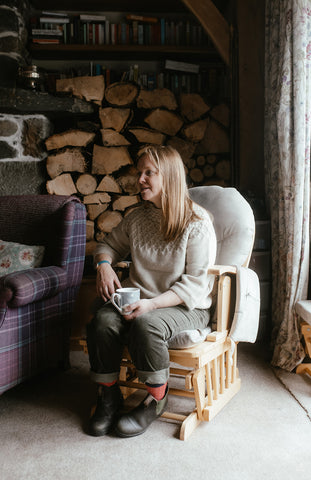 Helen Rebanks sits with a cup of tea in her living room, she is wearing a cream Elba Sweater and green cord trousers