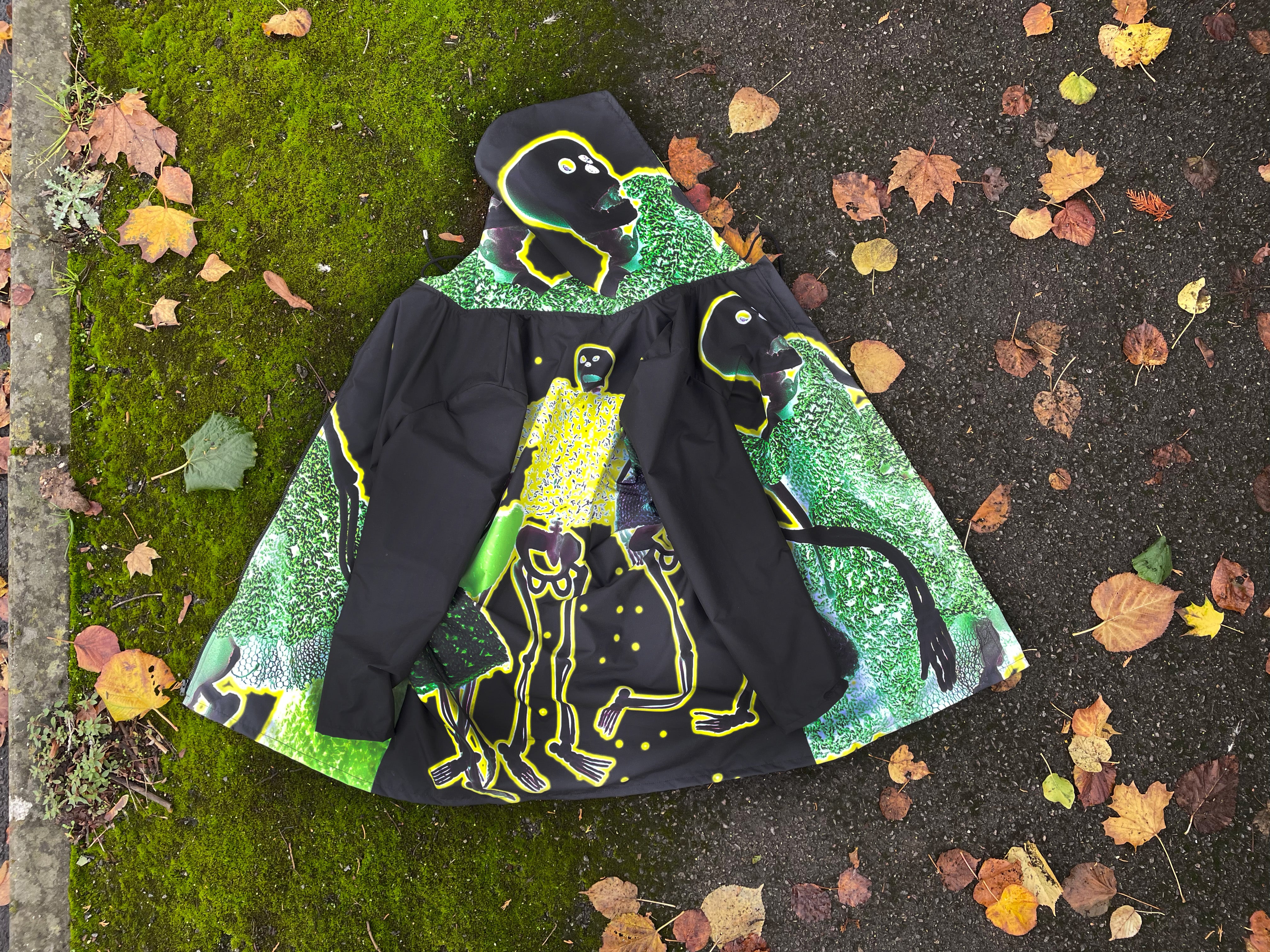 Black waterproof raincoat with skeletons print. On green moss and autumn leaves