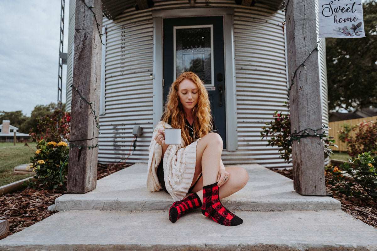 A woman sits on a front porch with a mug and wears fuzzy socks in black and red plaid