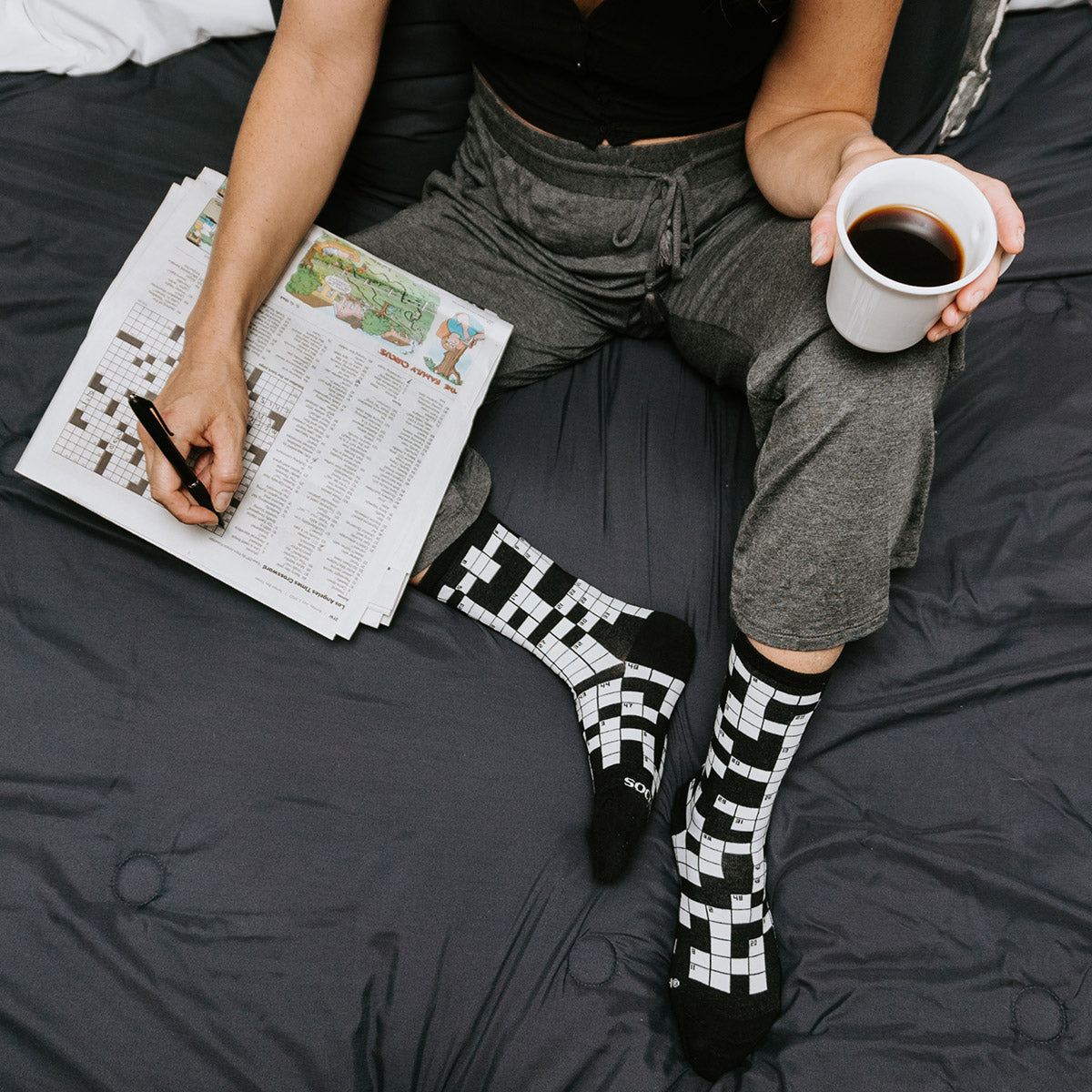 A woman wears crossword socks on a bed while doing a crossword puzzle and holding a cup of coffee